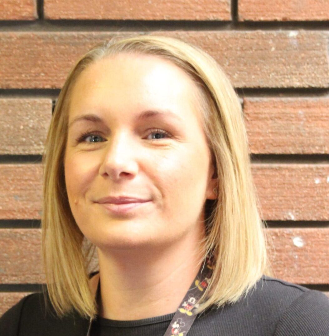 Smiling woman with straight blonde hair, wearing a black top and patterned lanyard, standing in front of a brick wall