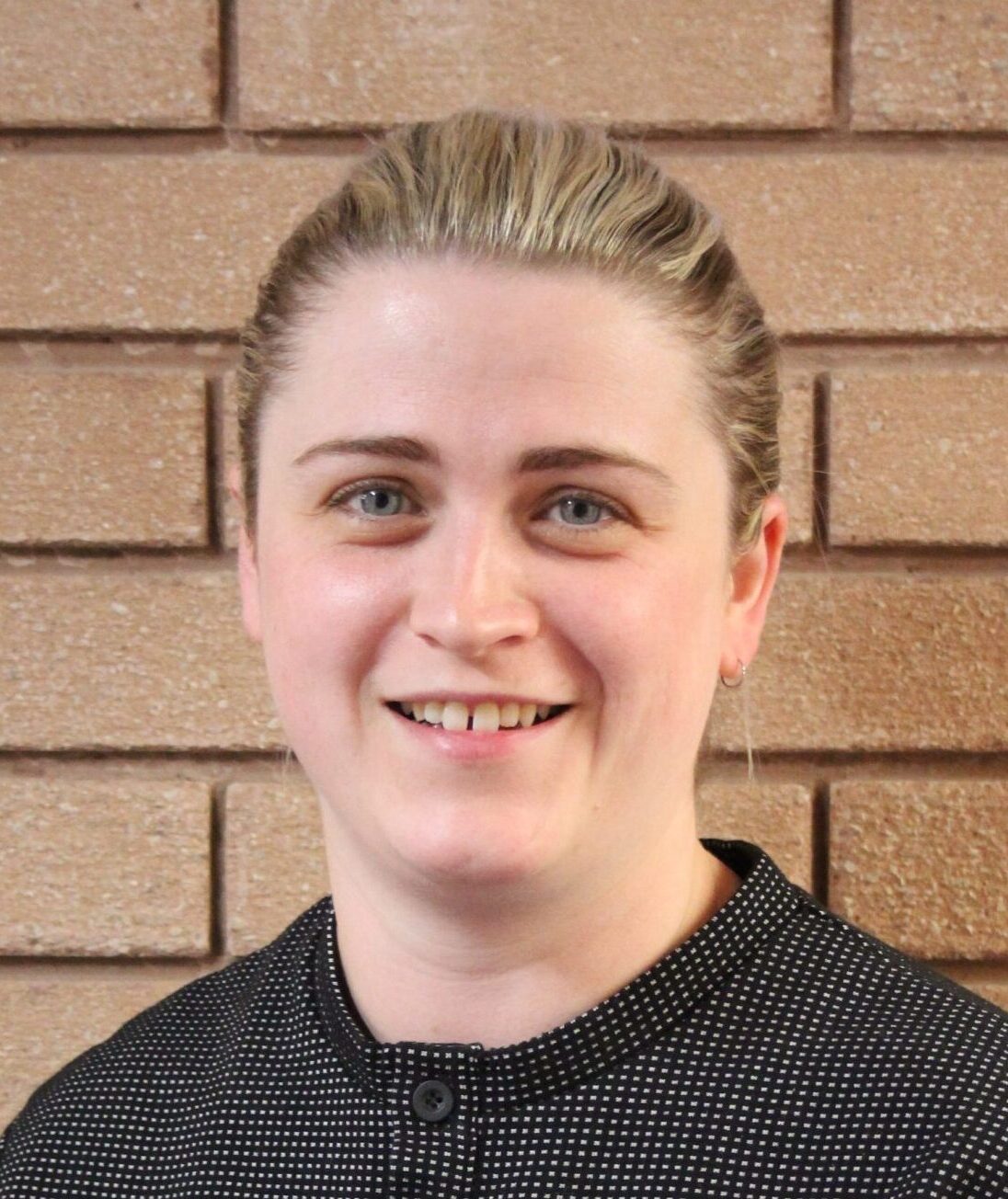 Smiling woman with light hair tied back, wearing a dark patterned top, standing in front of a brick wall