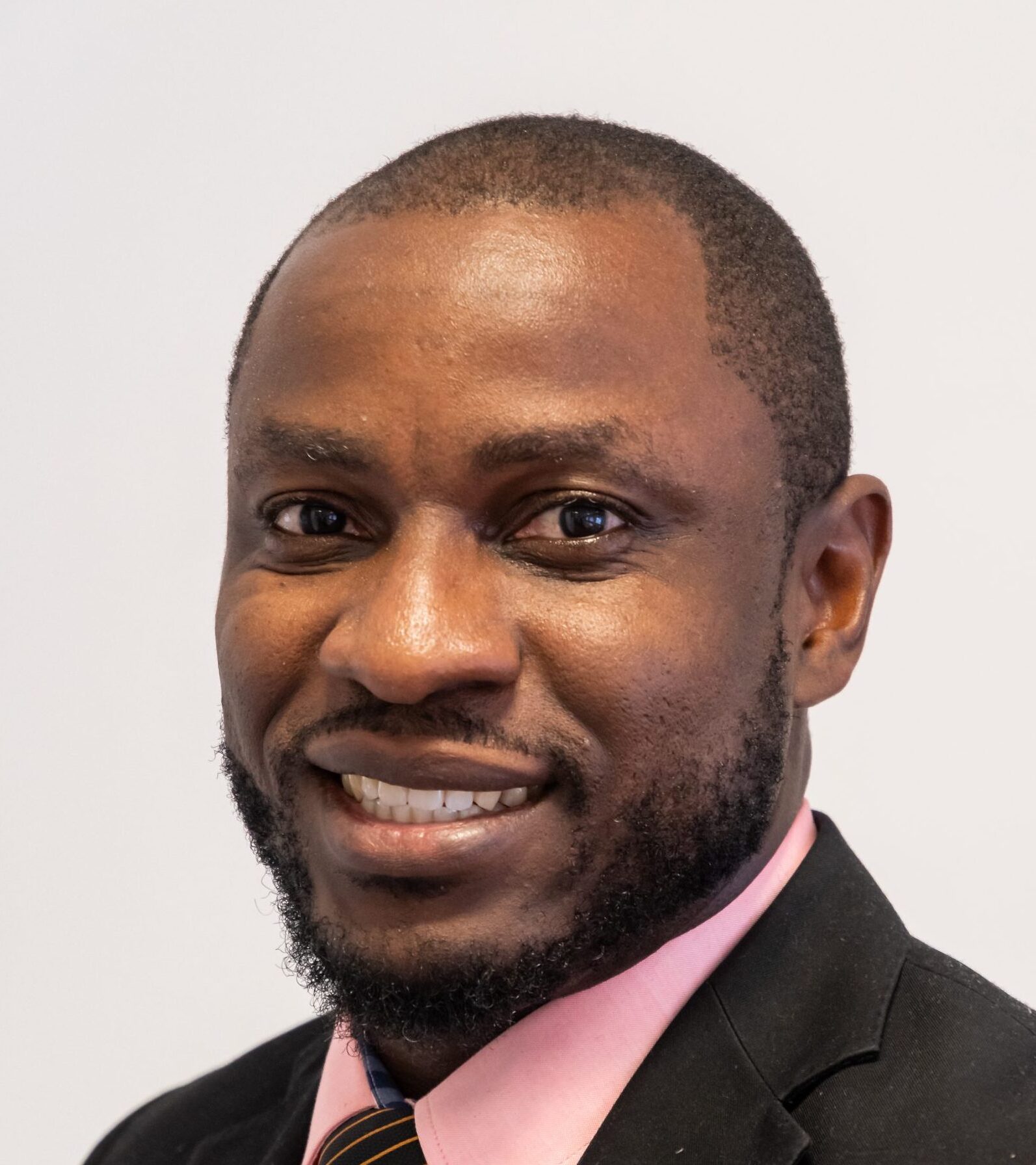 Smiling man wearing a suit jacket, pink shirt, and striped tie, posed against a plain light background