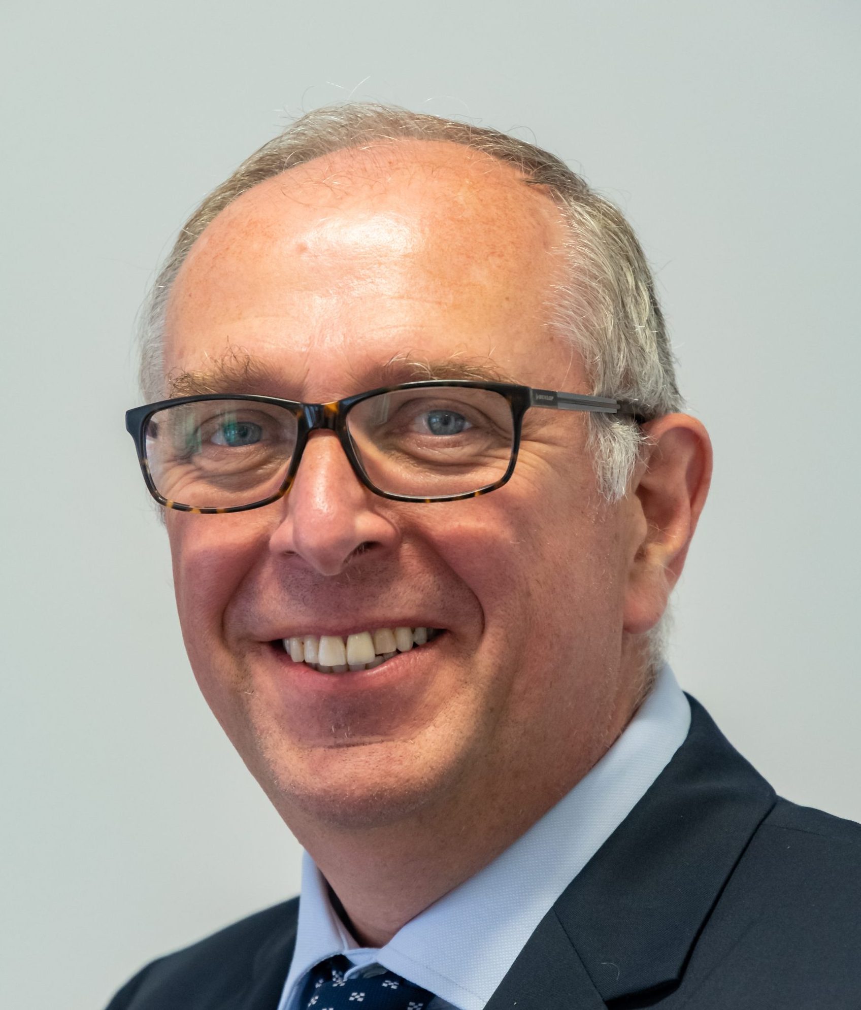 Smiling middle-aged man wearing glasses, a suit jacket, and a tie, posed against a plain light background