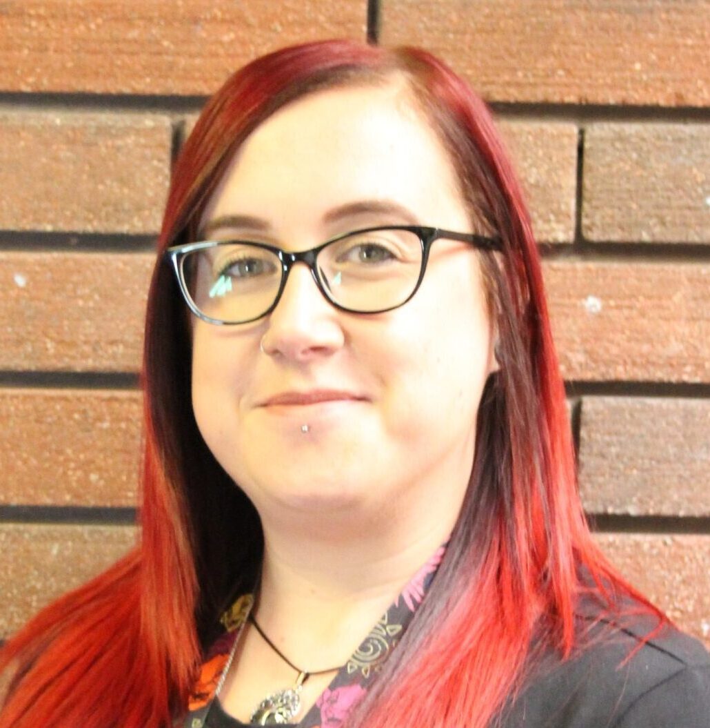 Smiling woman with straight red hair, wearing glasses and a black top, wearing a patterned lanyard, standing in front of a brick wall
