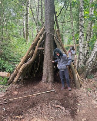 Young boy wearing a grey coat and grey Nike joggers, under a tree fort made with branches