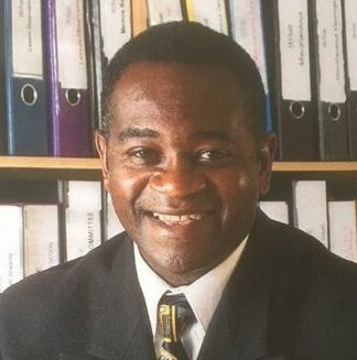 A man in a dark suit smiling while seated in front of a shelving unit filled with neatly organised ring binders