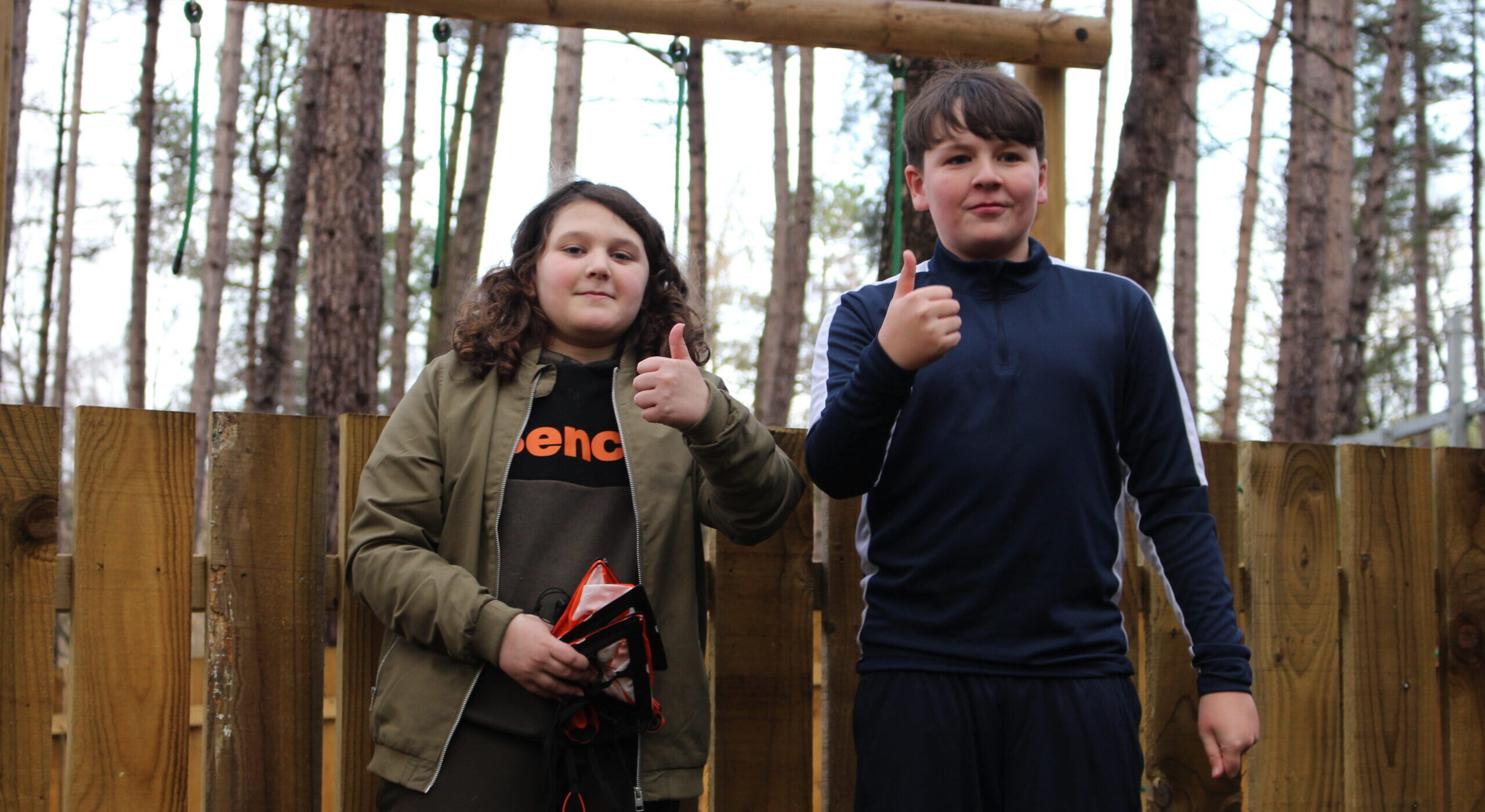 two young boys stood in the woods, smiling with their thumbs up