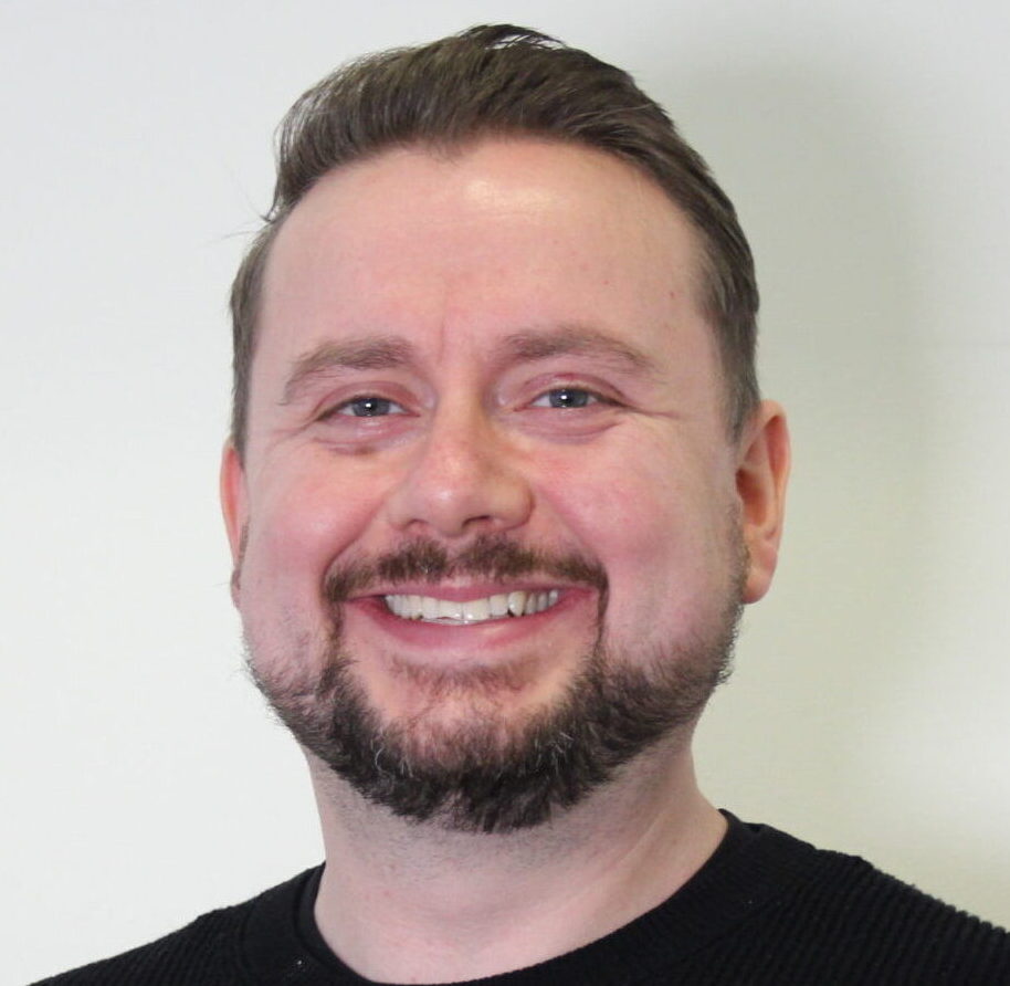 A smiling man with a beard standing indoors in front of a light-colored door, wearing a black top