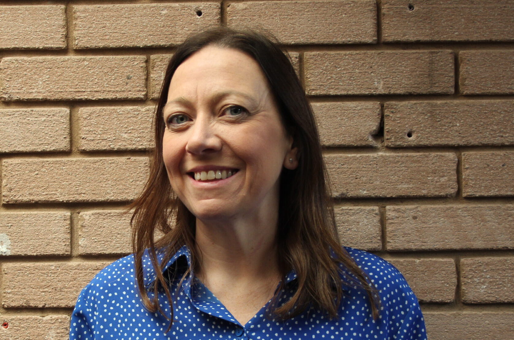 Brunette lady with a blue shirt, standing in front of a brick wall