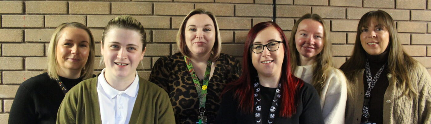 A group of six staff members standing side by side indoors against a brick wall, smiling at the camera. They are wearing smart-casual clothing and staff lanyards, representing a professional and welcoming team.