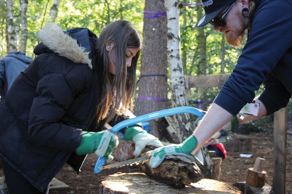 young girl with brown hair, black coat with fluffy hood and green gloves cutting a wooden log with a ginger haired man with a beard, black cap, blue jacket and green gloves