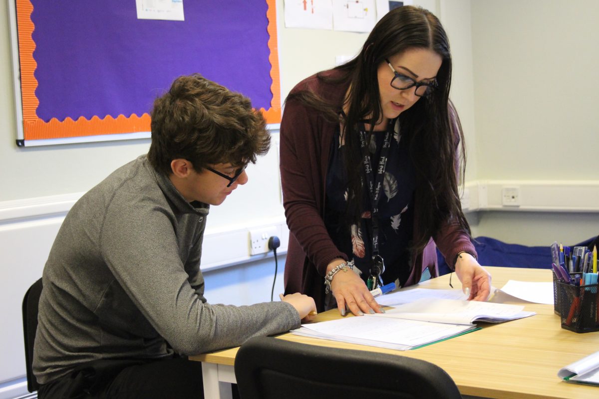 Brunette teacher wearing a floral dress and burgundy cardigan and brunette learner in a grey quarter zip top looking at a notebook in class.