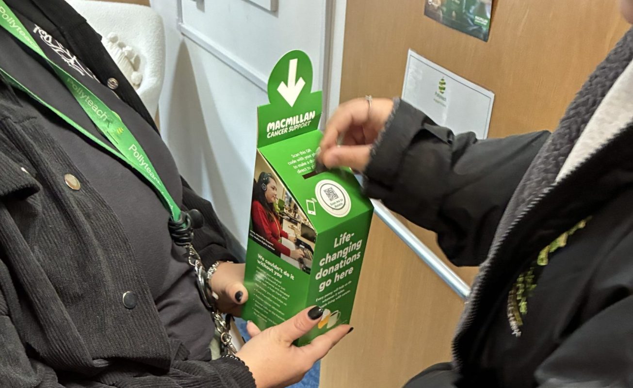 A girl wearing a black jacket puts money into a Macmillan donation cup while another girl in a brown jacket holds it.