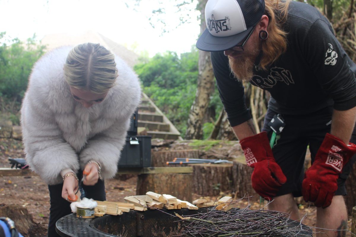 Blonde girl wearing a fluffy jacket attempting to light a fire with male ginger teacher