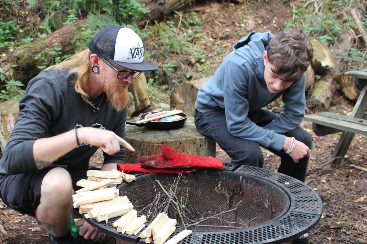 a ginger bearded male teacher and brunette boy ina blue hoodie learning about firepit safety.